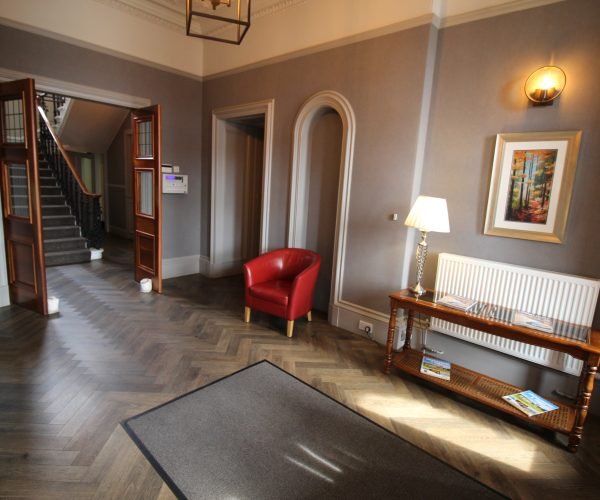 A hallway with a wooden floor and a red chair leading to a hotel room.